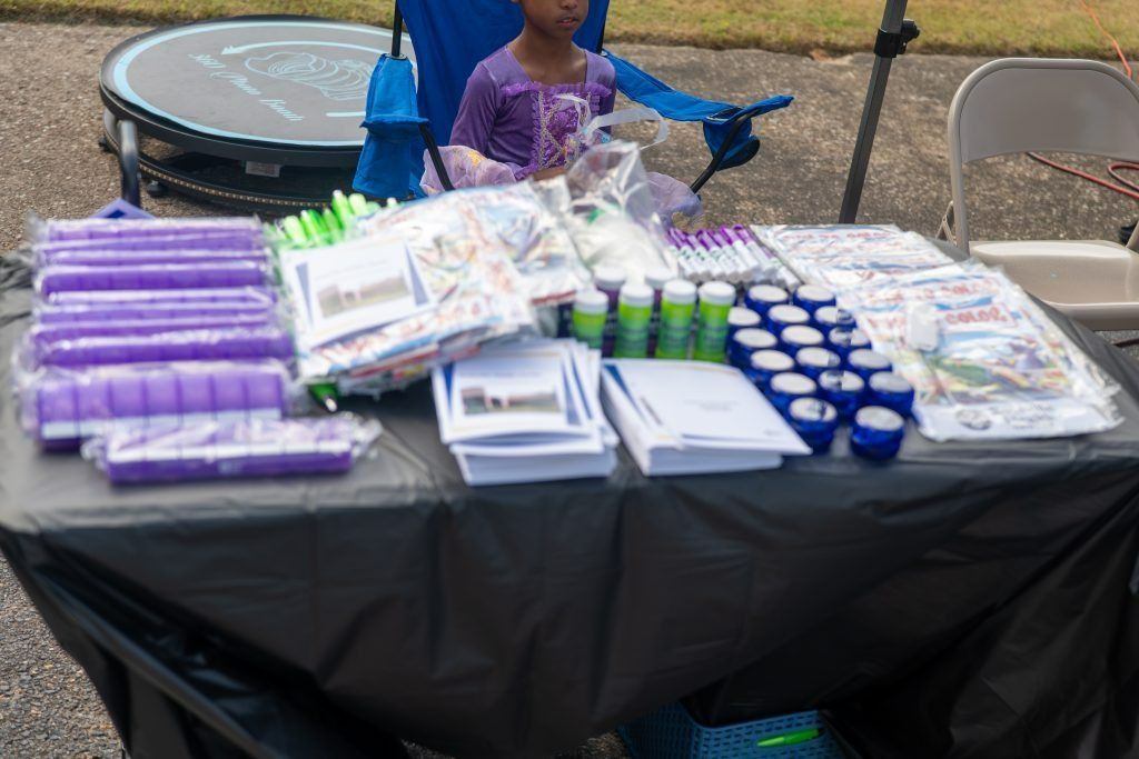 Table with neatly arranged packaged items and small blue cups, with a child standing behind it outdoors.