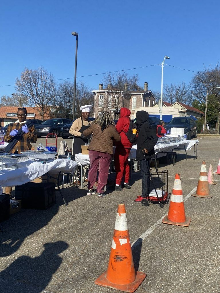 People standing at outdoor tables beside traffic cones and folding tables in a sunny parking lot.