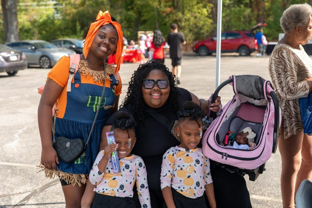 Three people pose outdoors, with two children in front and a pink stroller at right.