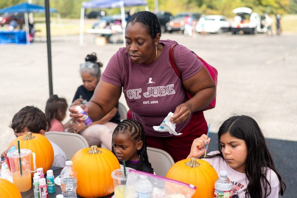 Children painting orange pumpkins at an outdoor table while an adult assists them