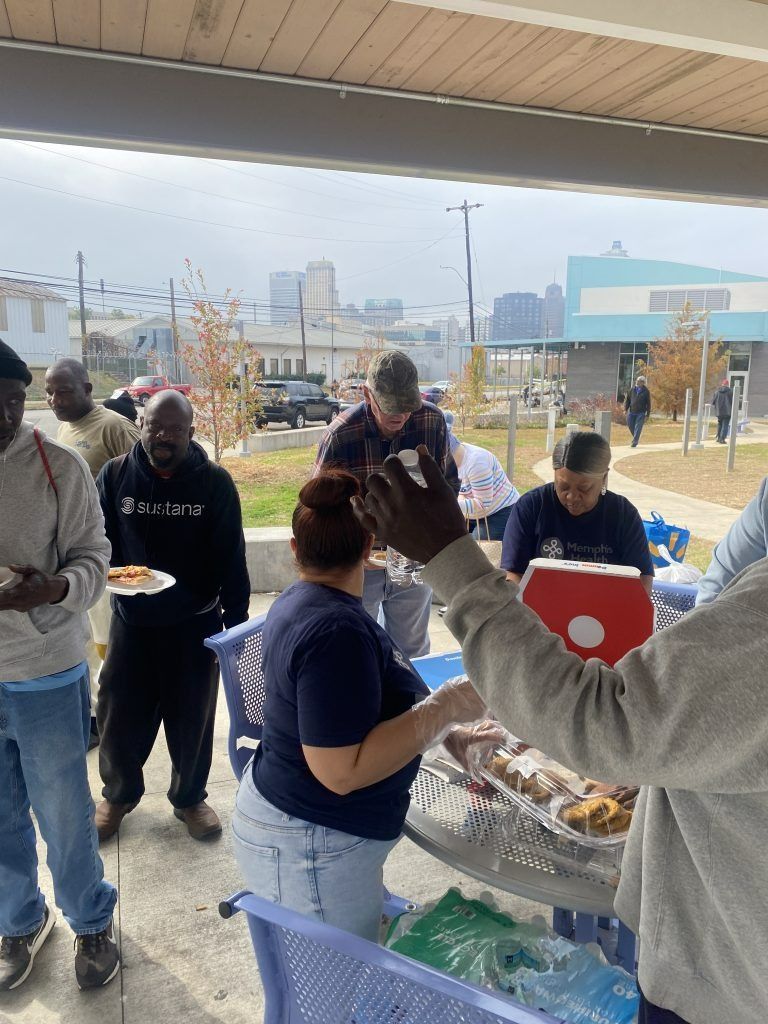 People gathered outdoors around a table with trays of food and drinks under a covered patio.