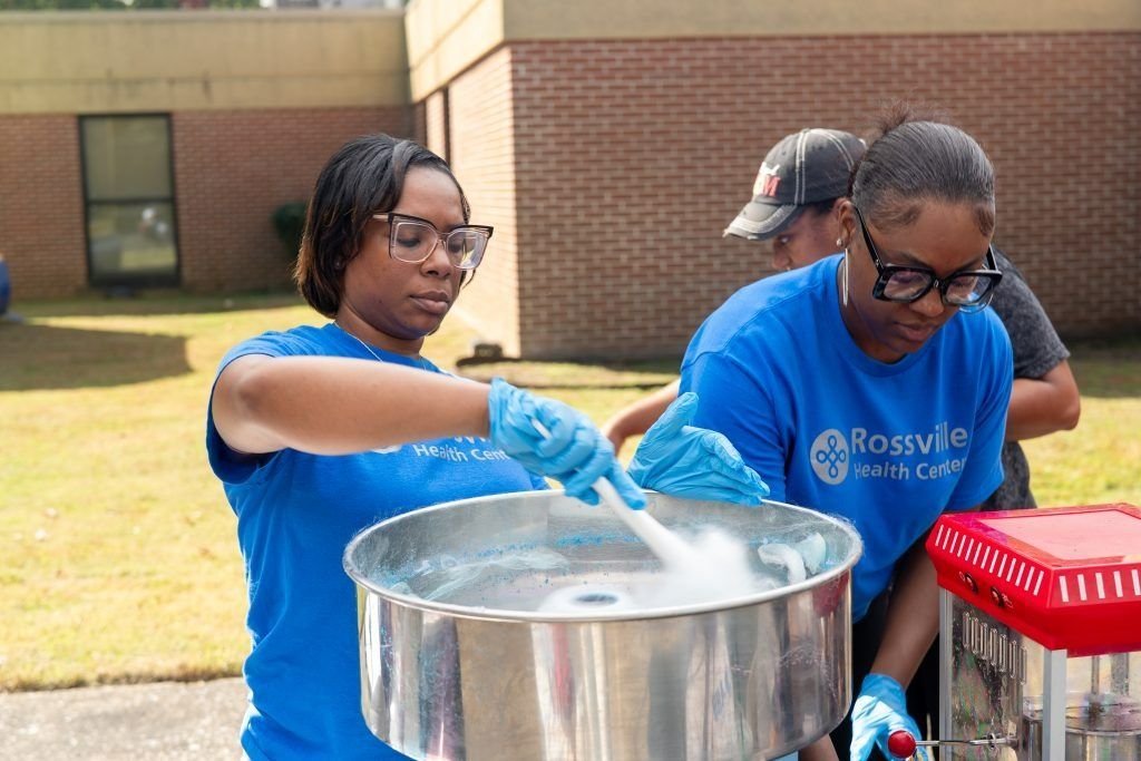 Two people in blue shirts stirring a large metal pot outdoors at a community event