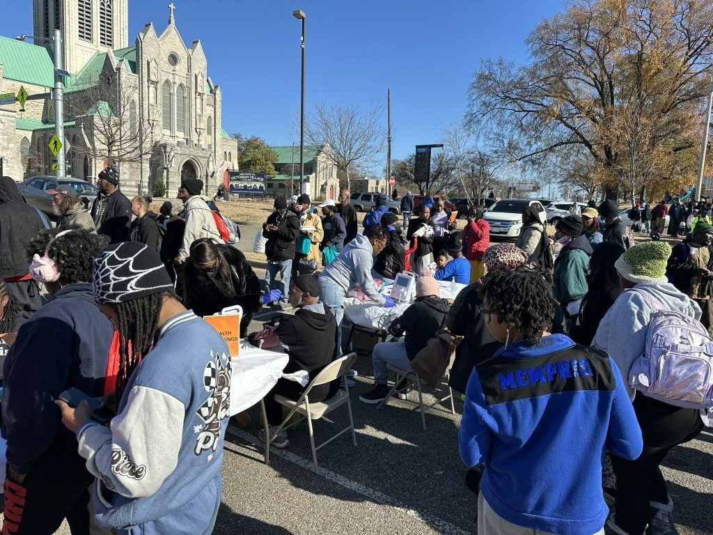 Crowd gathered outdoors near a stone building, many seated with signs at a public event.