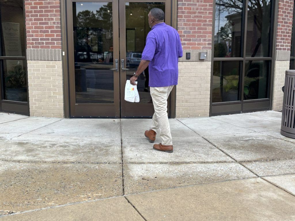 Man in purple shirt walking into a brick building, carrying a white bag