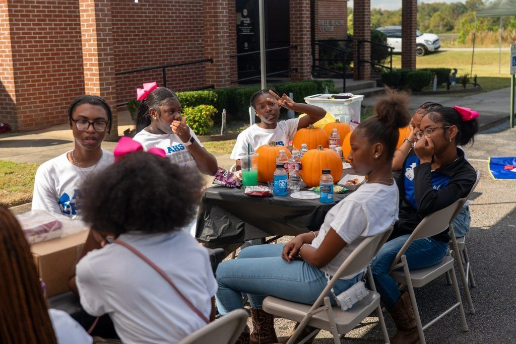 People seated around a table with pumpkins at an outdoor gathering by a brick building