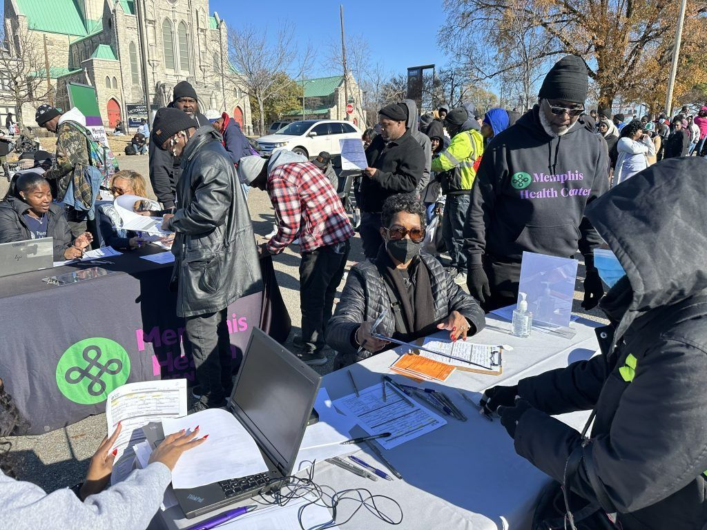 People gathered at an outdoor protest, with tables, laptops, signs, and a city skyline in the background.
