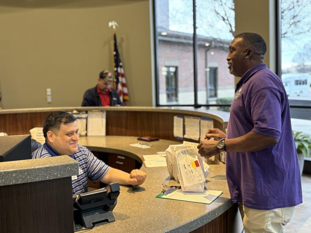 Two people talking at a courthouse counter, with an American flag and large window in the background.