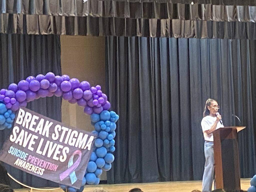 Speaker at podium beside purple balloon arch and sign reading “Break Stigma Save Lives” on stage