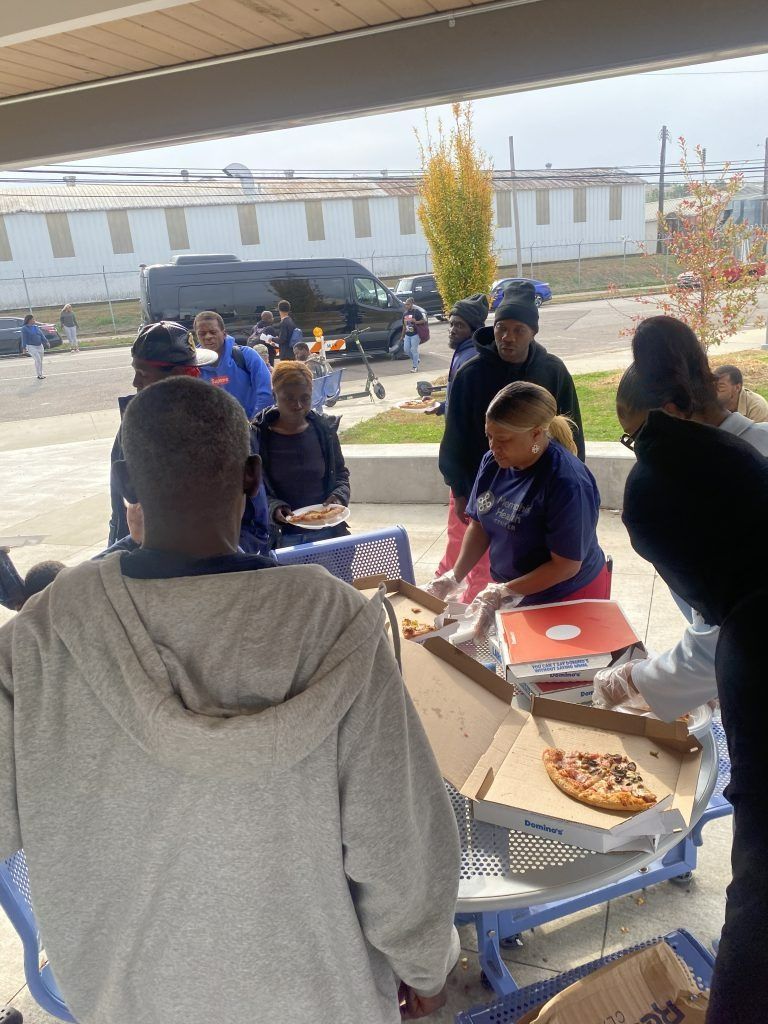 People gathered around a blue picnic table outdoors, serving food on a sunny day.