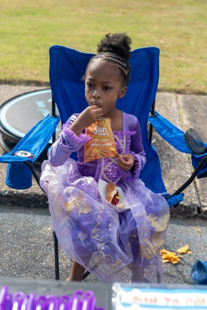 Child in a purple princess dress sitting on a blue folding chair and eating a snack outdoors.