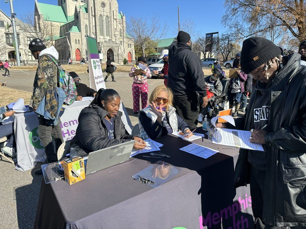 Outdoors at a table, two people assist others with papers during a community event near a stone building.