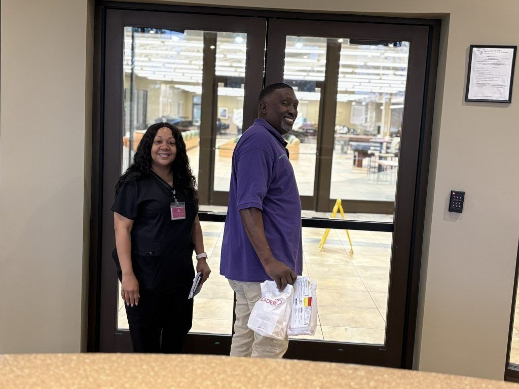 Two people stand by glass doors in a lobby, one holding plastic bags.