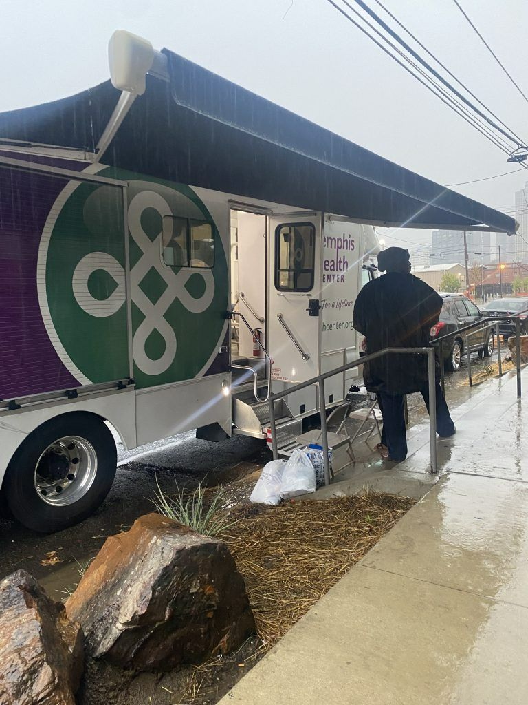Food truck with green logo parked by a sidewalk in the rain; person stands outside near a table