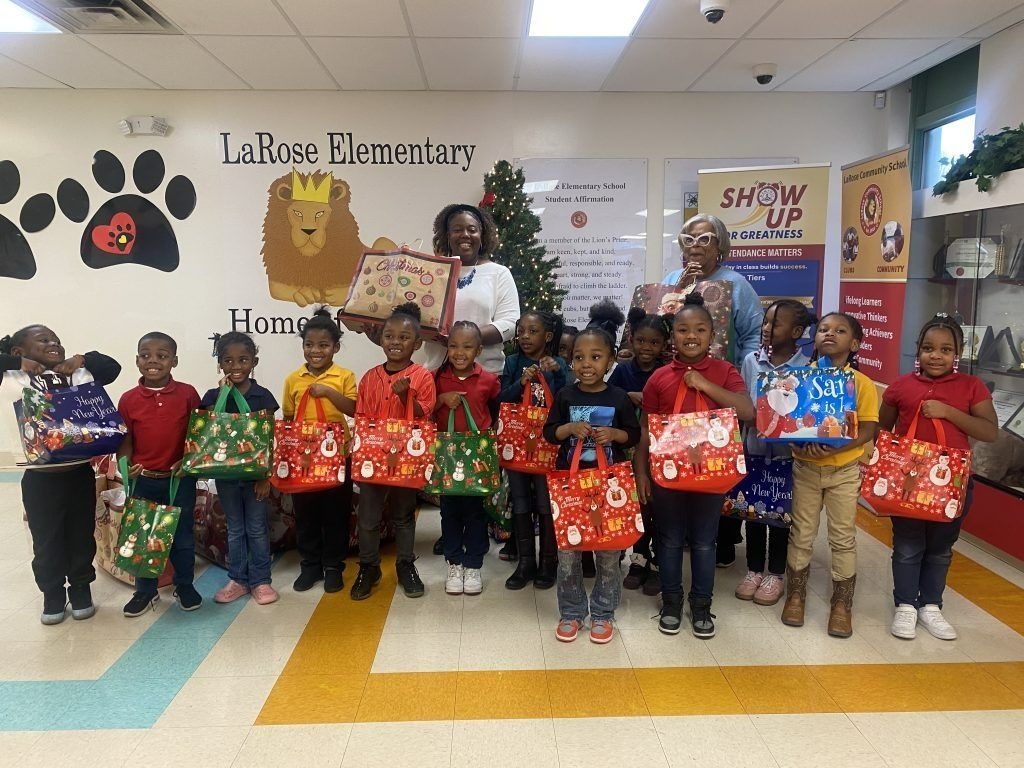 Children holding holiday gift bags with a teacher at an elementary school celebration, standing in a classroom.