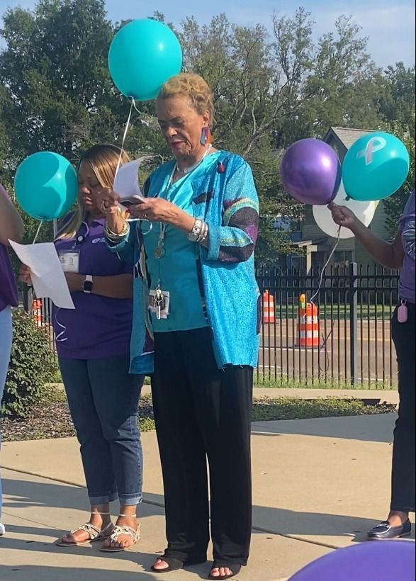 Woman in blue jacket speaking outdoors, holding papers, with balloons nearby.