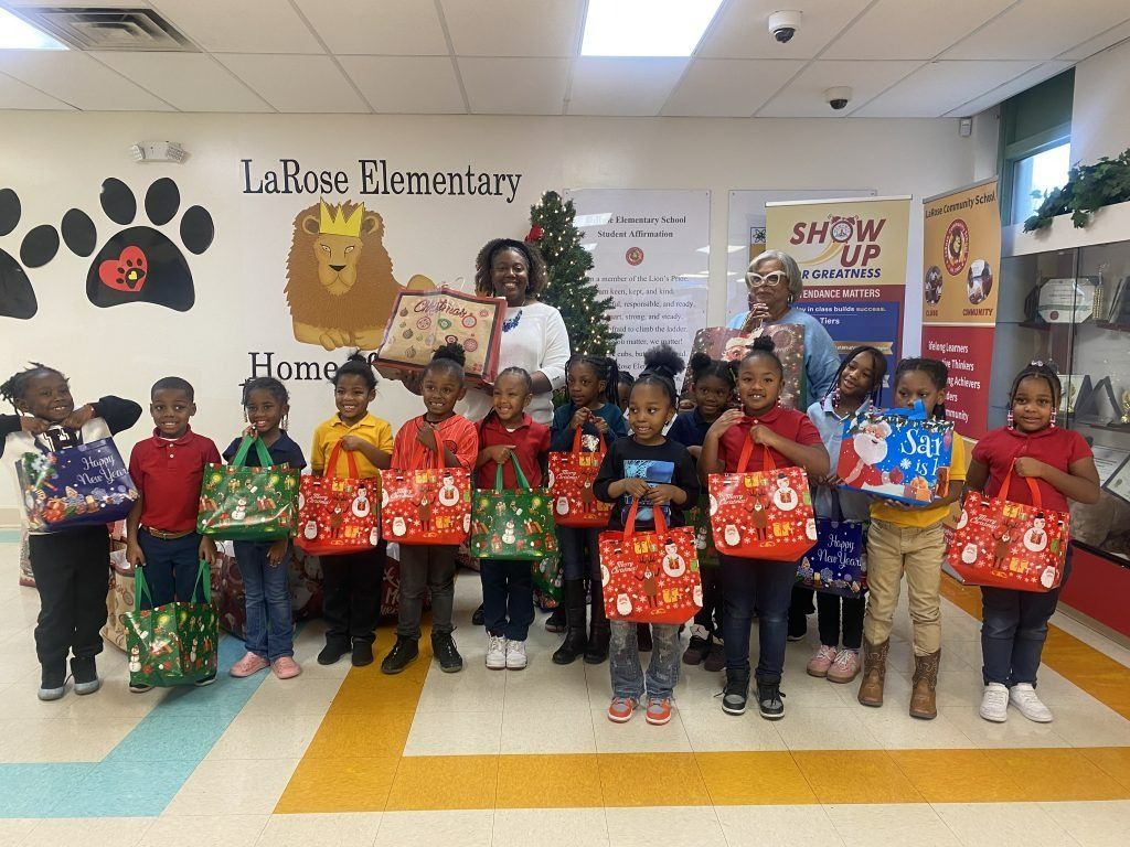 Children holding holiday gift bags in a school classroom, standing in a row under festive decorations.