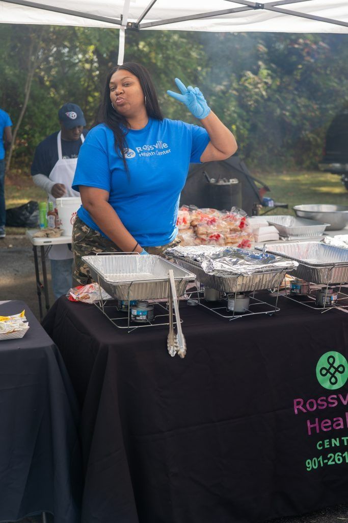 Volunteer in blue shirt and gloves serving food at an outdoor health event under a white tent.