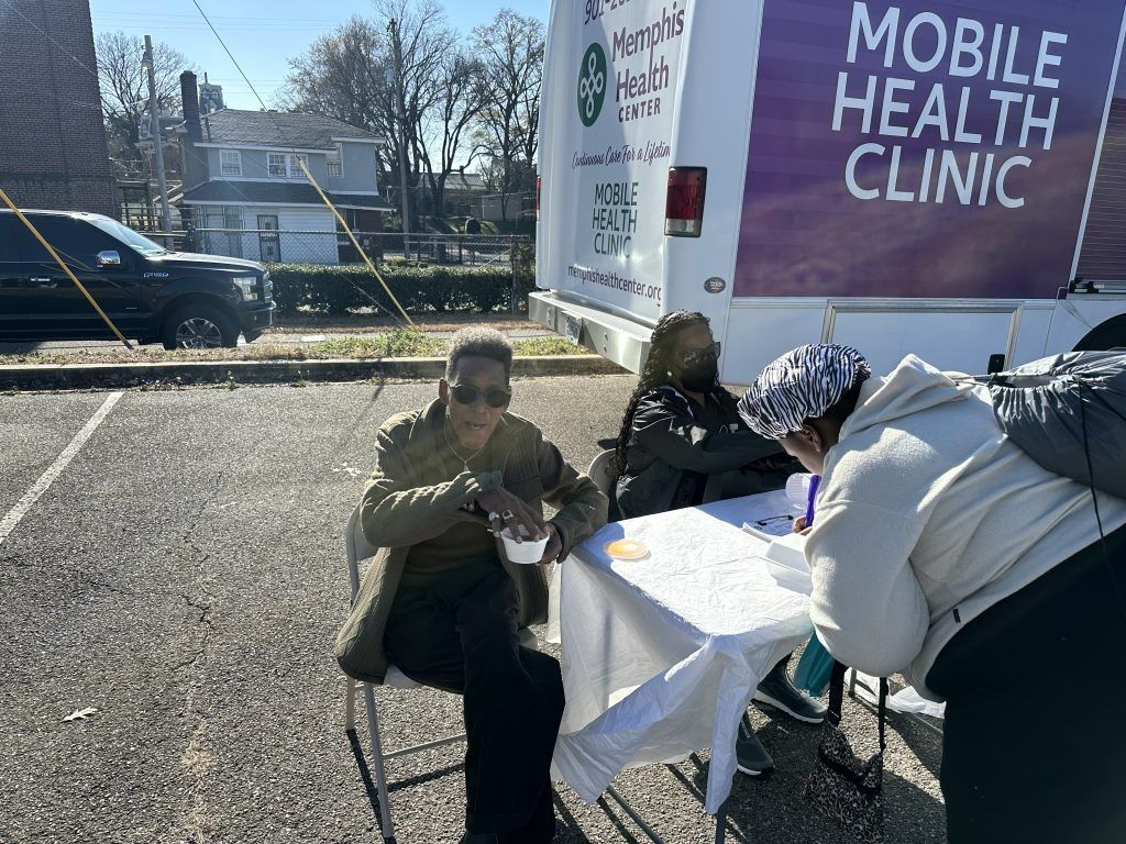 People seated outside a mobile health clinic tent on a sunny day, with a white table and banner visible.