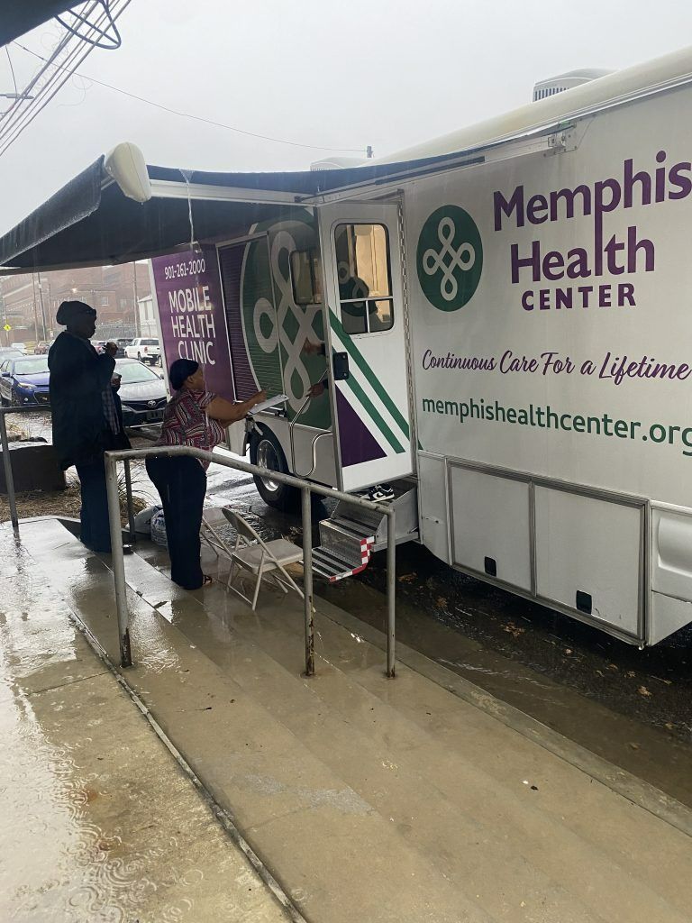Memphis Health Center mobile clinic at rainy sidewalk, with staff assisting a person under an awning