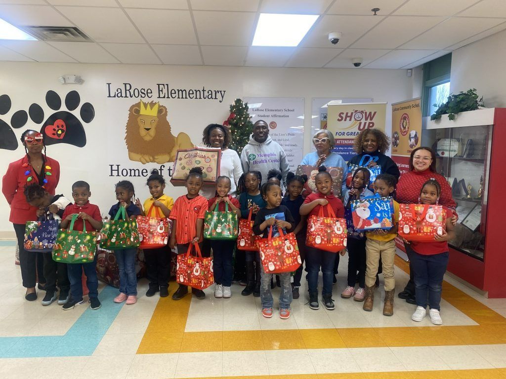 Group of children and adults holding wrapped gifts in a school hallway with festive holiday decorations