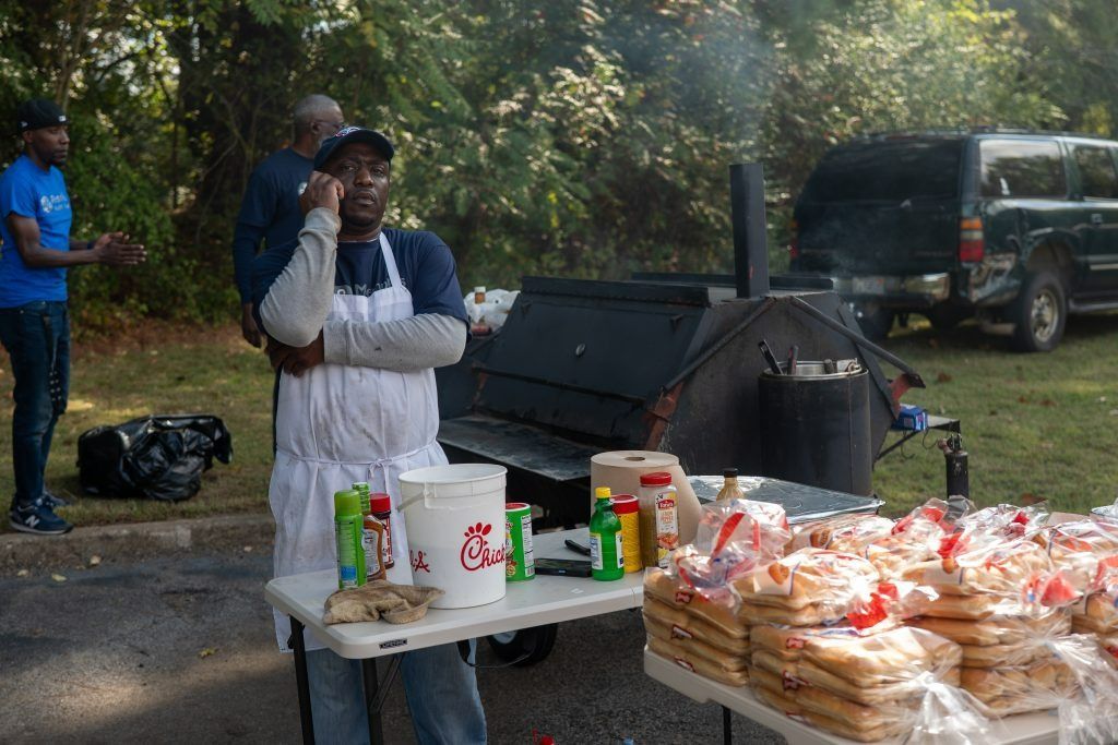 Person at an outdoor table with drinks and bread near camping gear and a parked vehicle