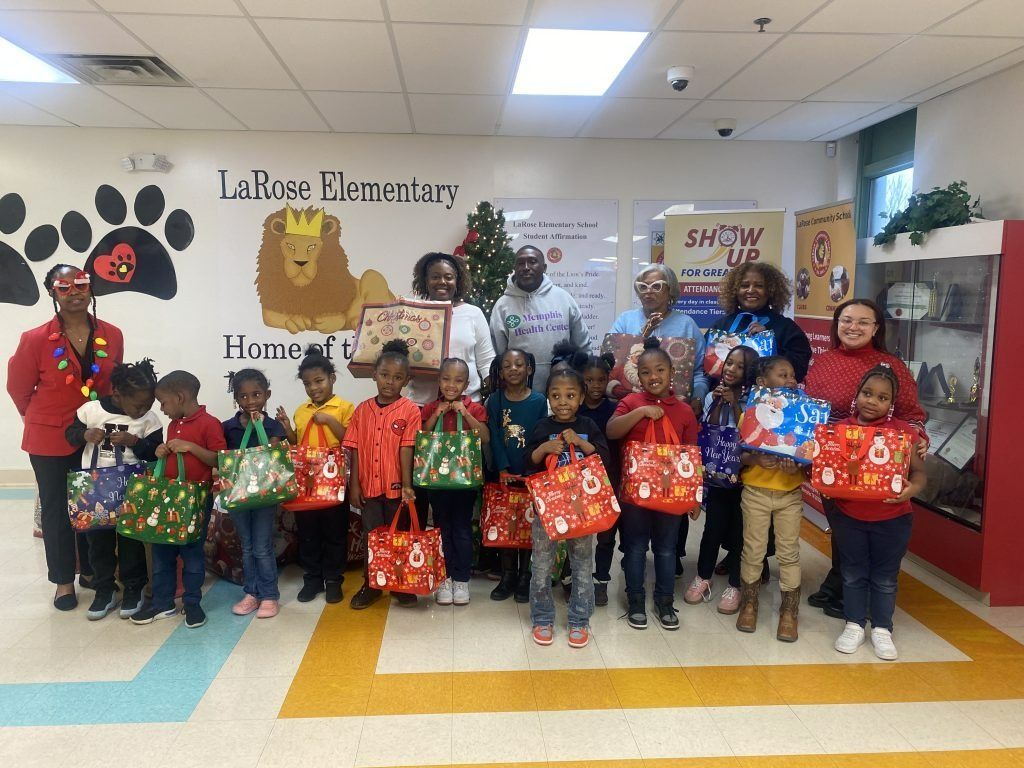 Group of children holding holiday gift bags with adults in a school hallway decorated with paw prints and banners.