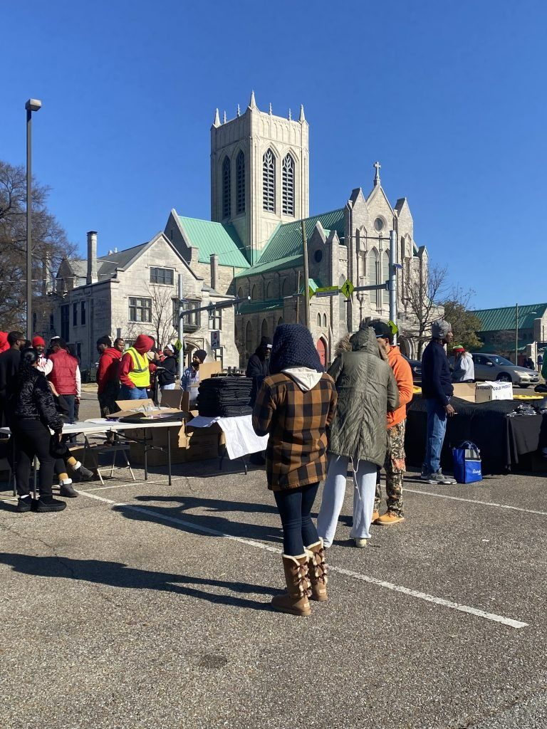 People gathered at an outdoor market in front of a stone church with a tall tower on a sunny day