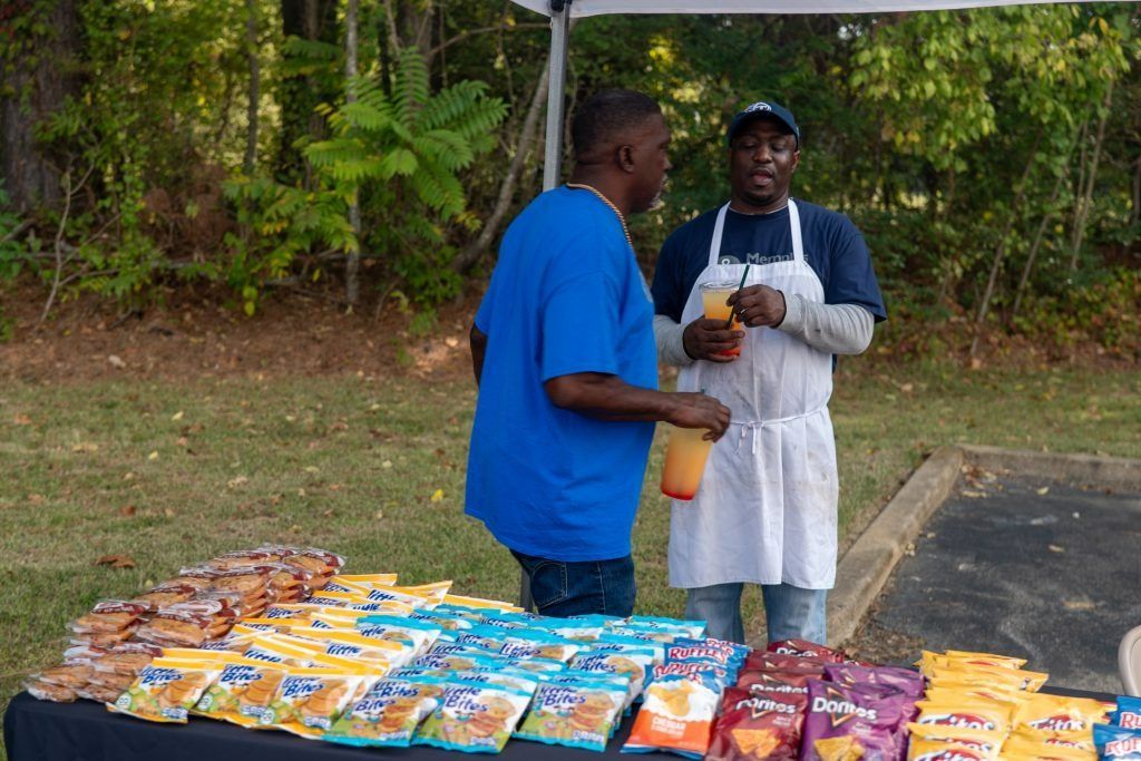 Two people at an outdoor market table with colorful packaged snacks displayed in front of them