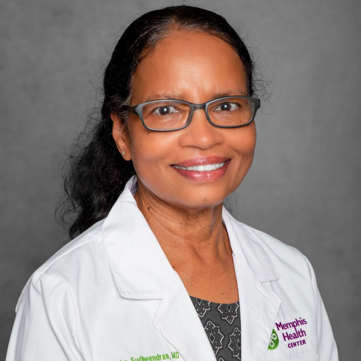 Woman in a white lab coat and glasses smiling against a gray background