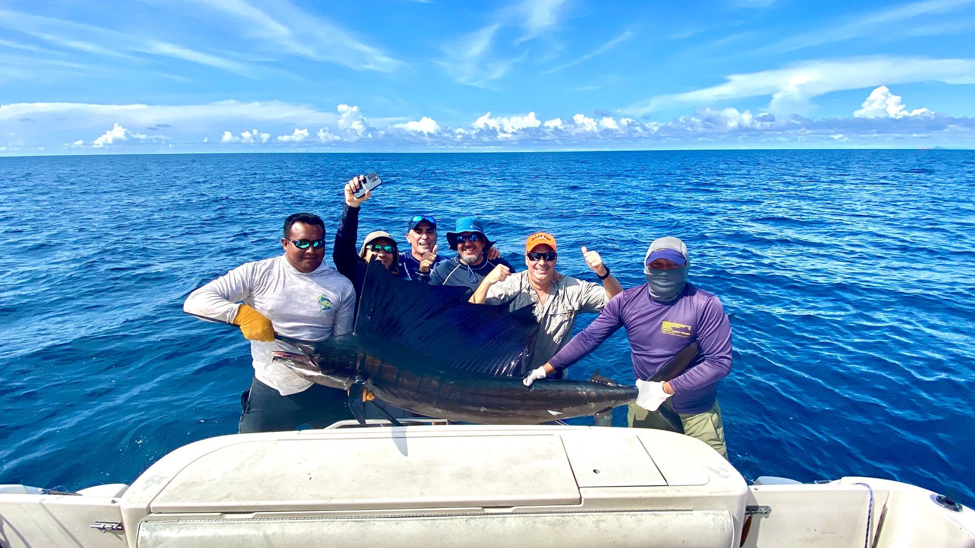 People on a boat pose with a large fish they caught, blue water and sky in background.