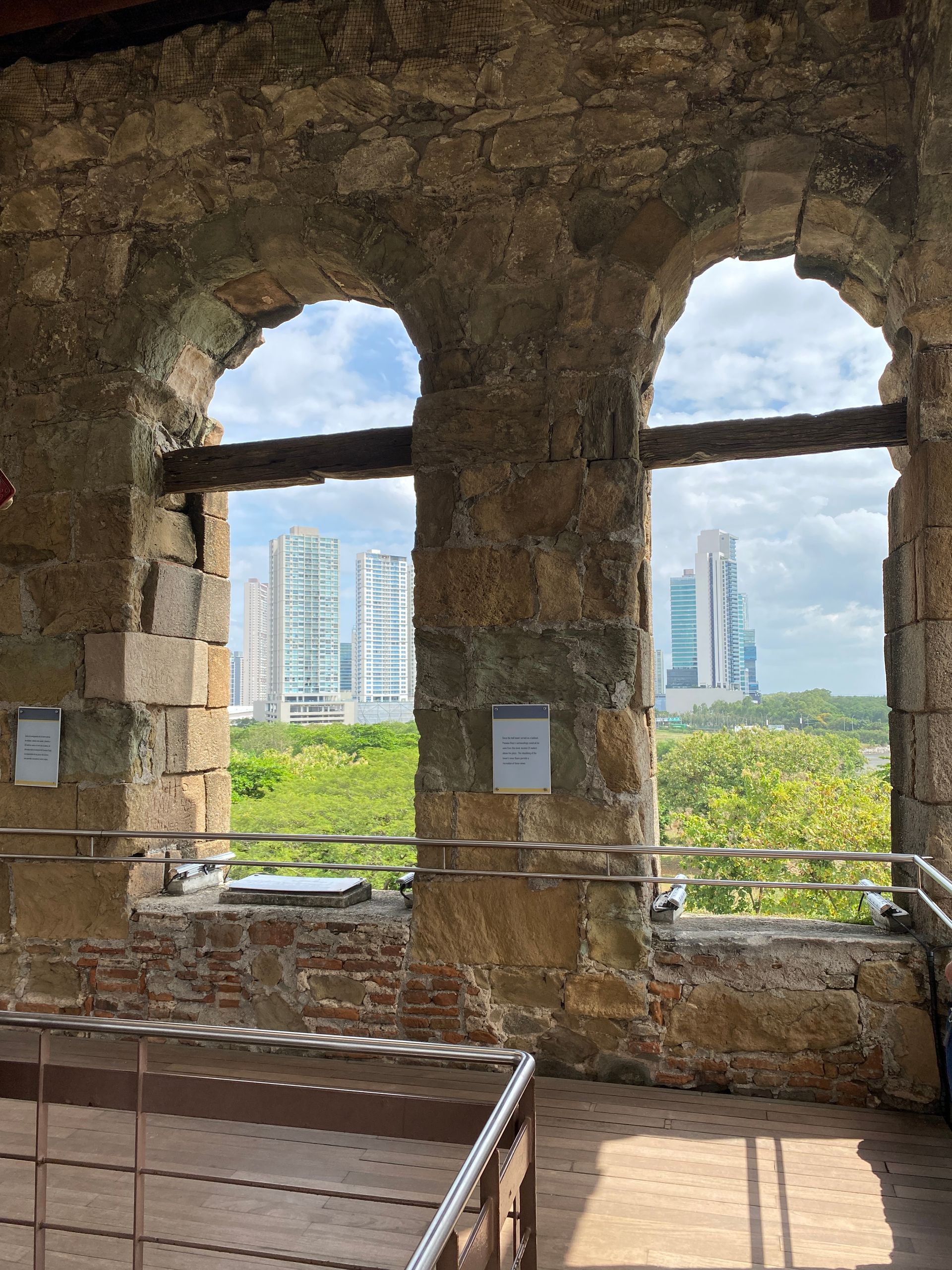 Interior view through two arched windows in an old stone building. Modern city buildings and green trees are visible outside.