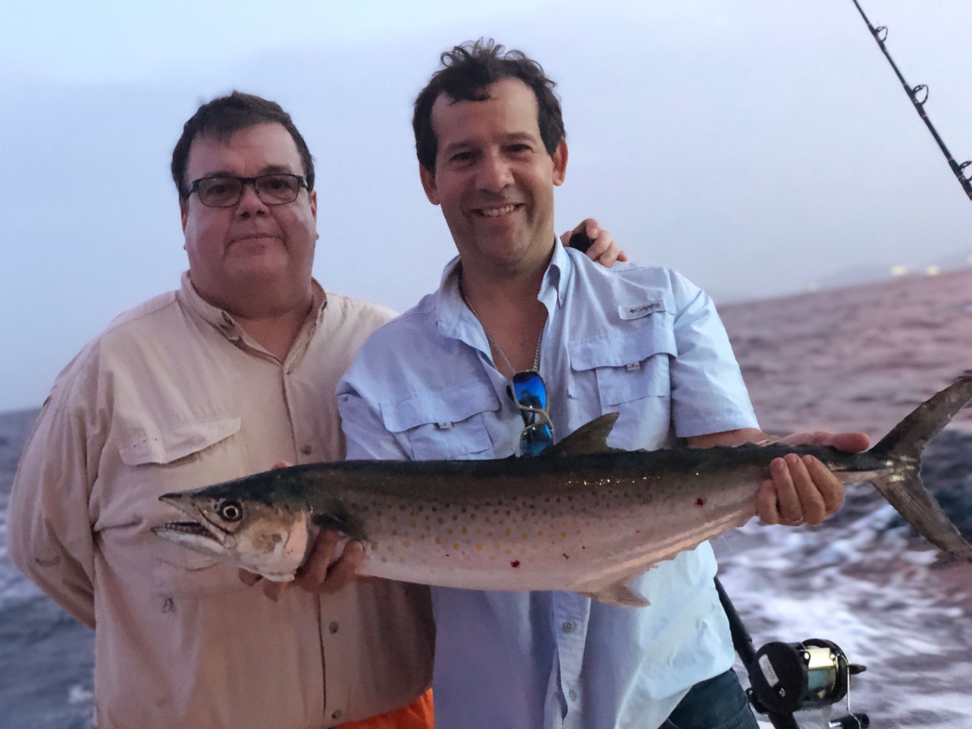 Two men on a boat, holding a large fish they caught. One man smiles, the other looks at the camera. Ocean background.