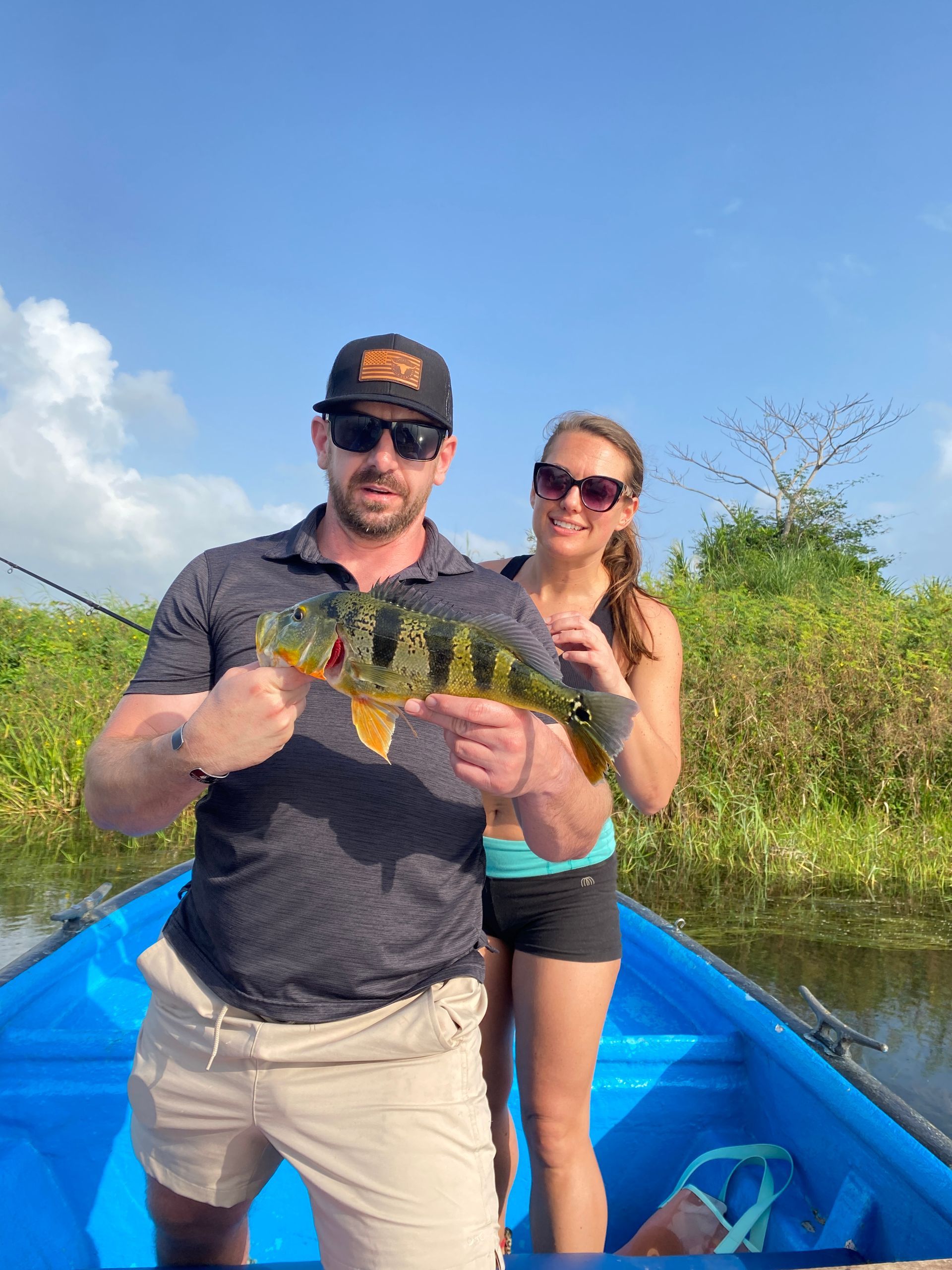 Man holding fish in a blue boat, woman standing behind him. Green vegetation and a bright blue sky are in the background.