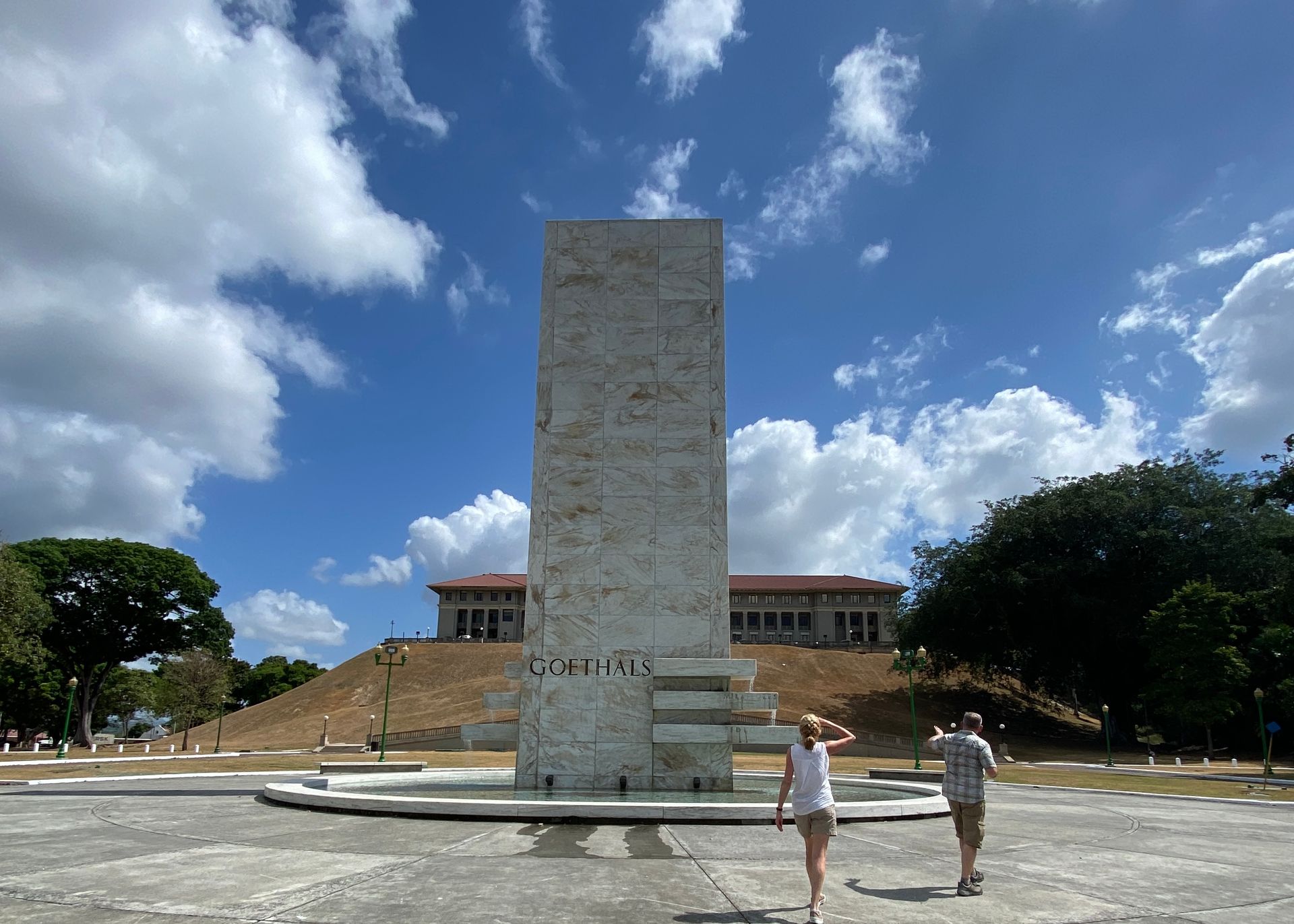 Tall white monument to Mayor General George W. Goethals Army Corps of Engineers in a plaza, two people walking, blue sky with clouds, Panama Canal Administration Building on a hill in background.