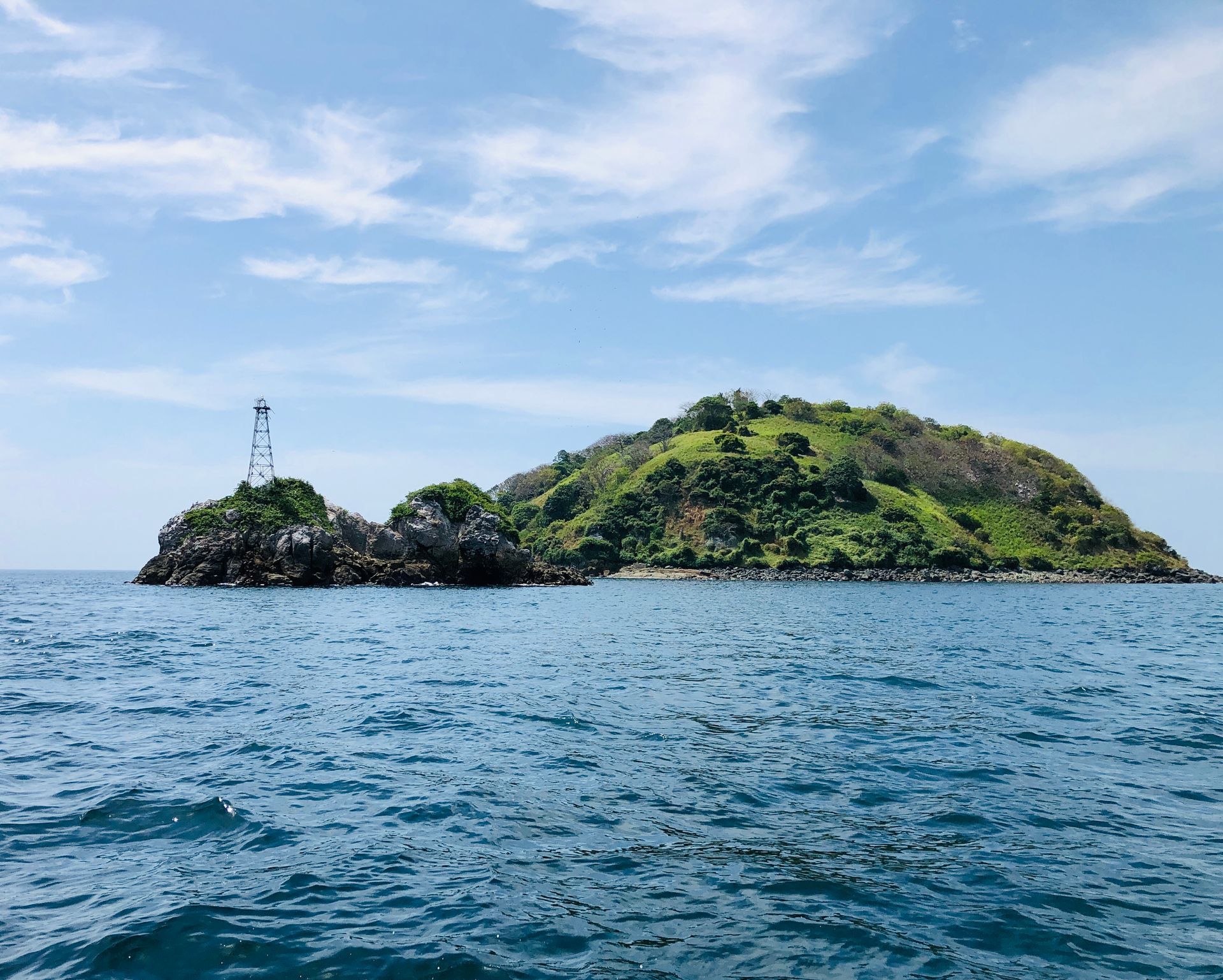 Island in turquoise water under a blue sky. Lighthouse on a small rocky islet, lush green hill behind.