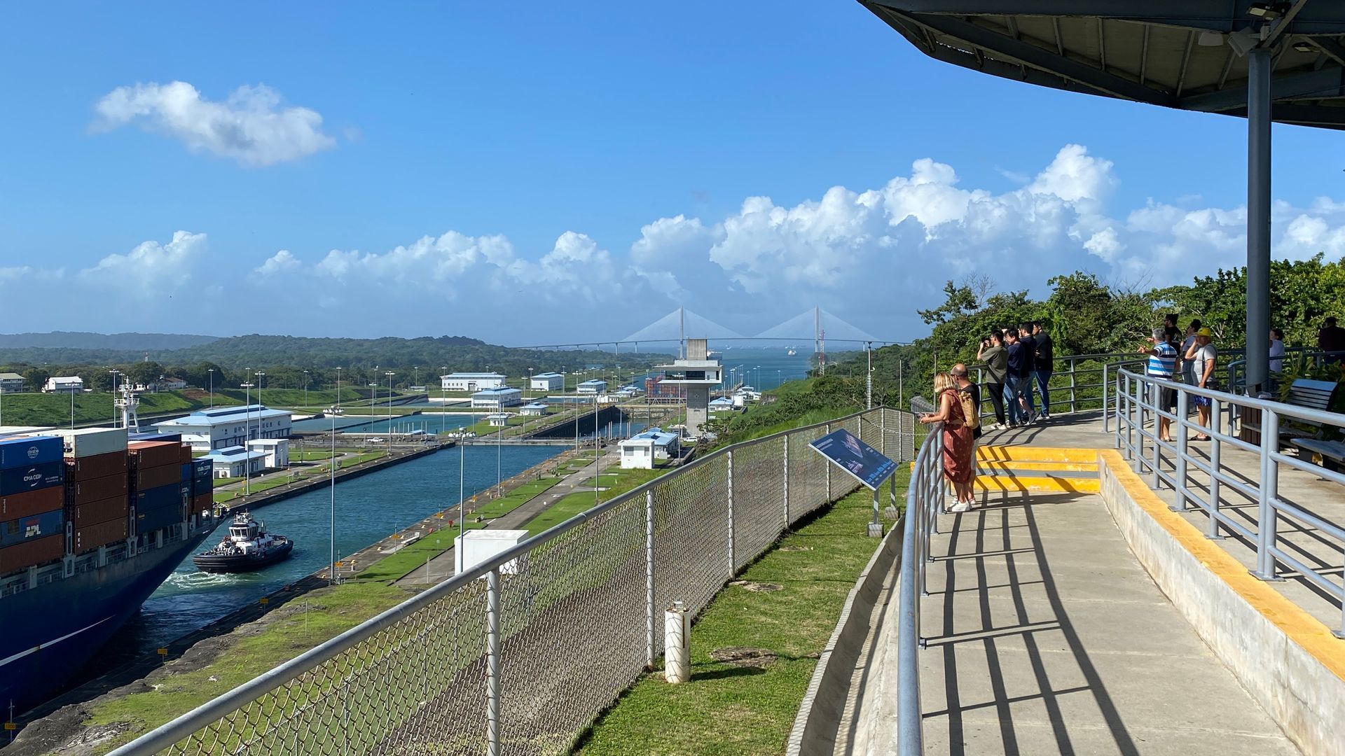 Container ship in canal, people on walkway. Blue sky, bridge in distance, green landscape.