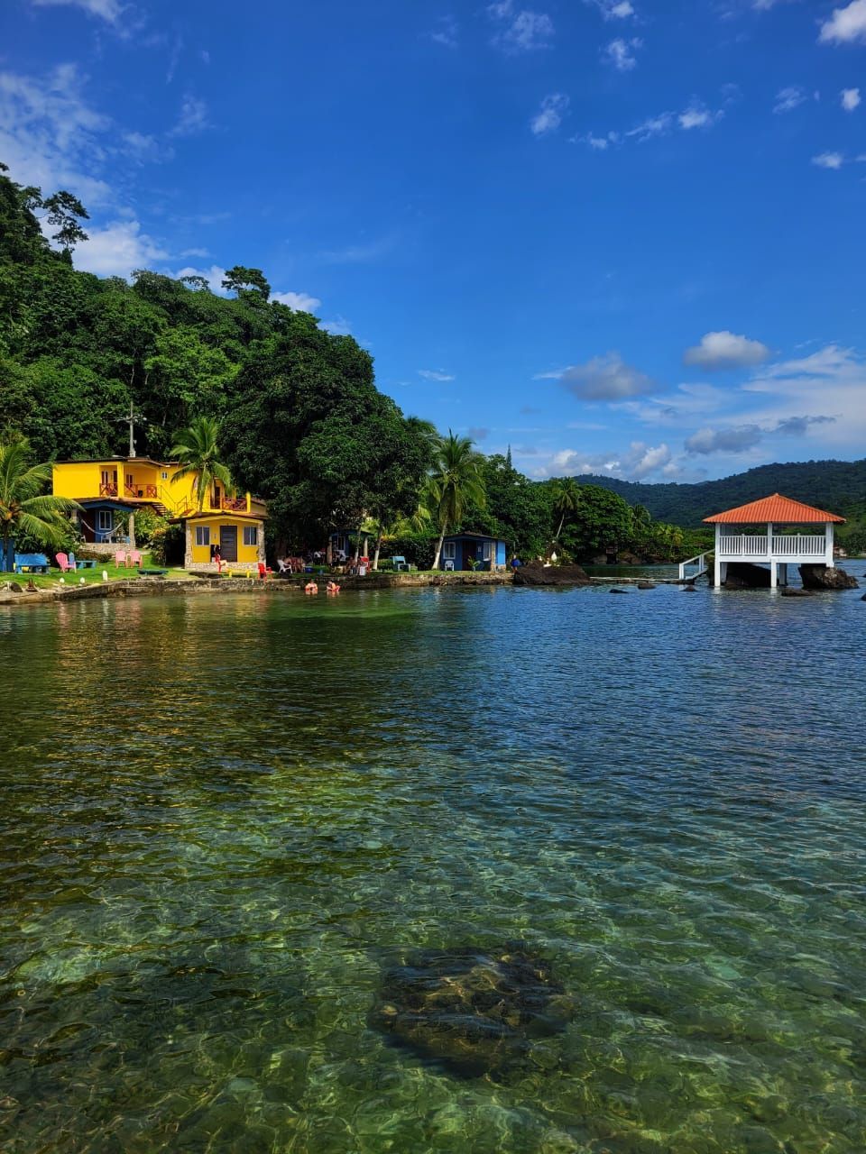 Coastal scene with clear water, a yellow building, and a white gazebo on pilings; lush green hills under a bright blue sky.