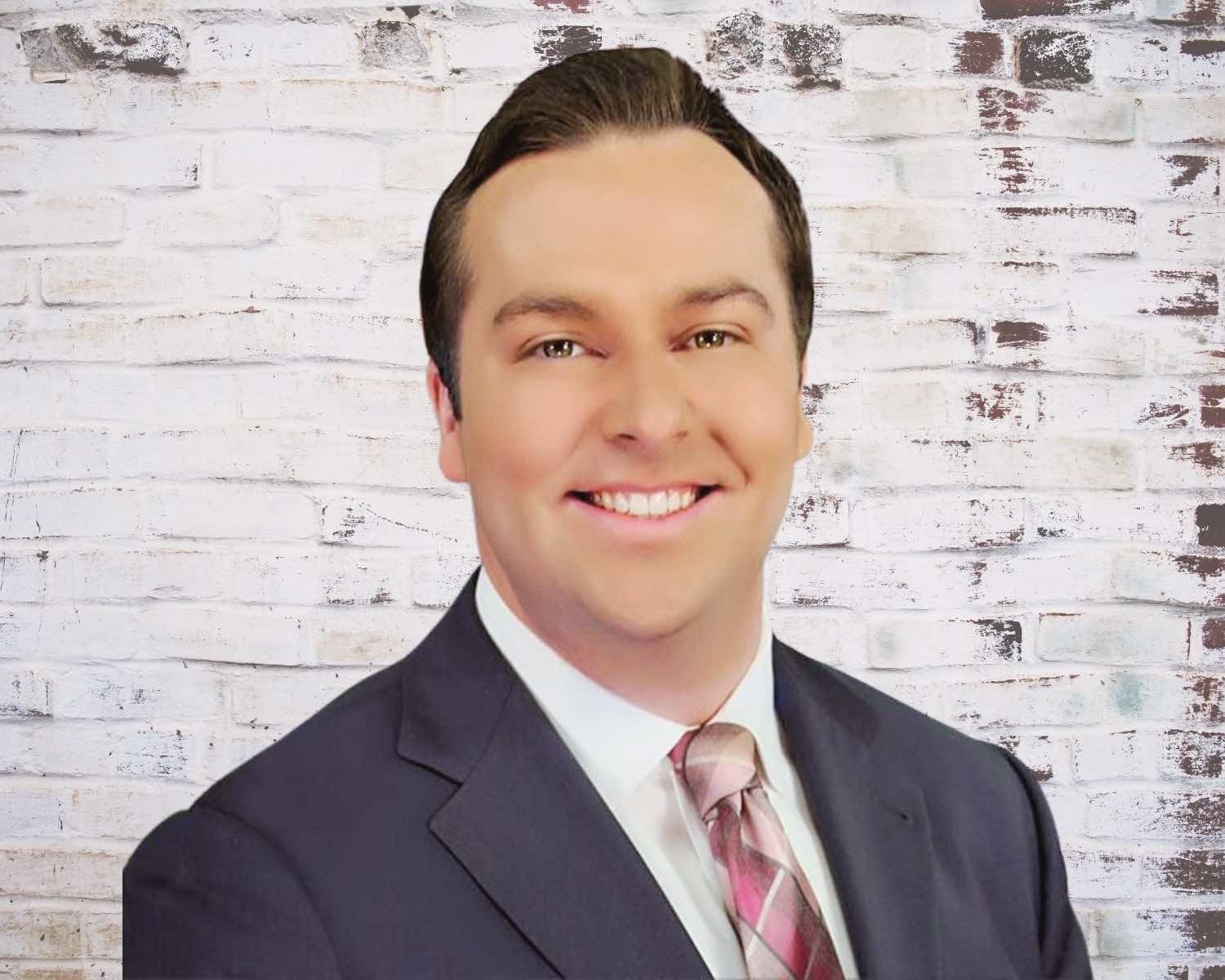 Man in navy suit and patterned tie smiling in front of a white brick wall.