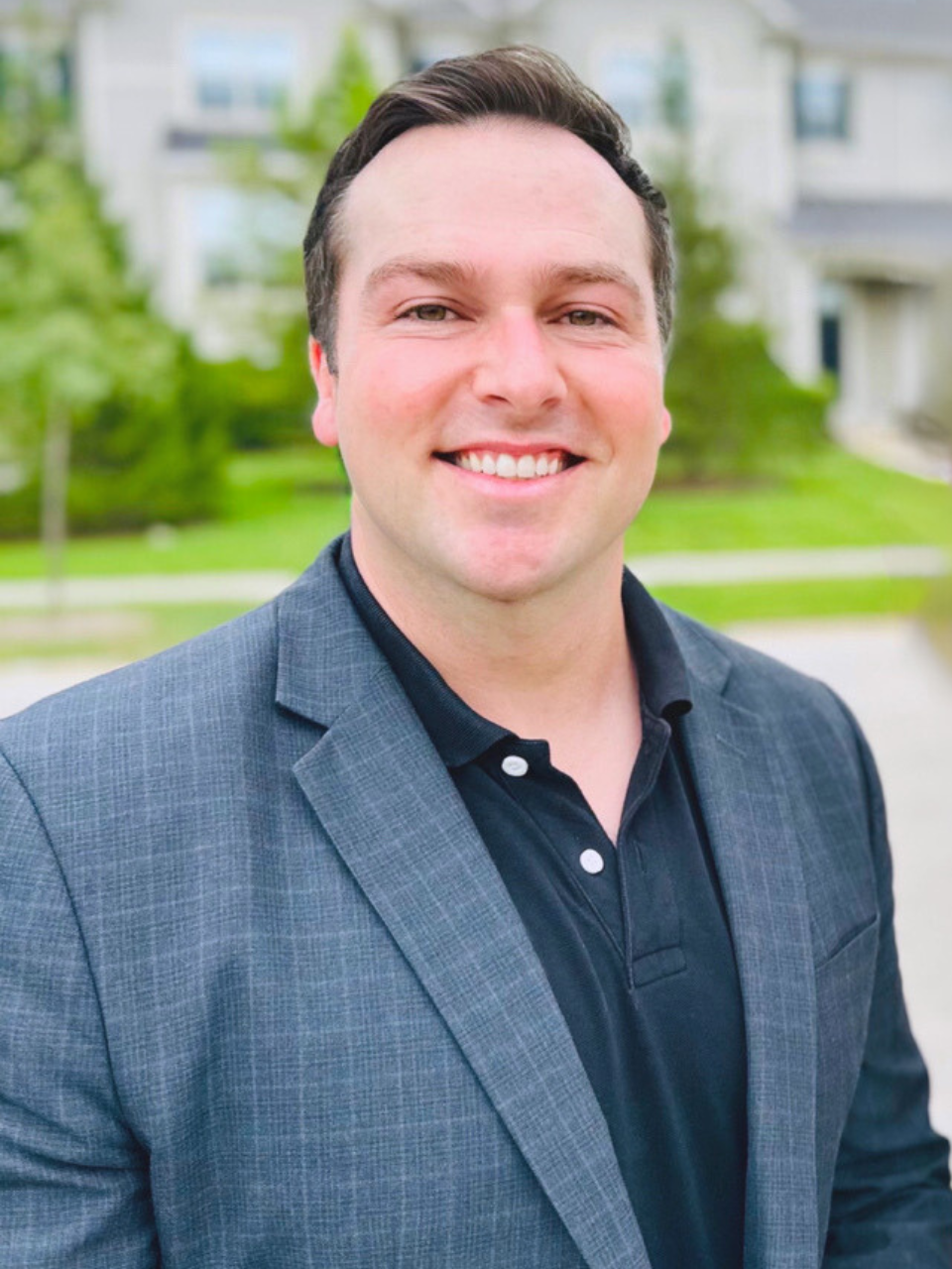 Man in gray blazer and black polo smiles outdoors near a house.