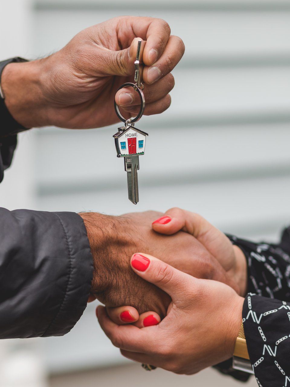 Hands exchanging house keys, one shaking another's hand. Keys have a small house-shaped fob.