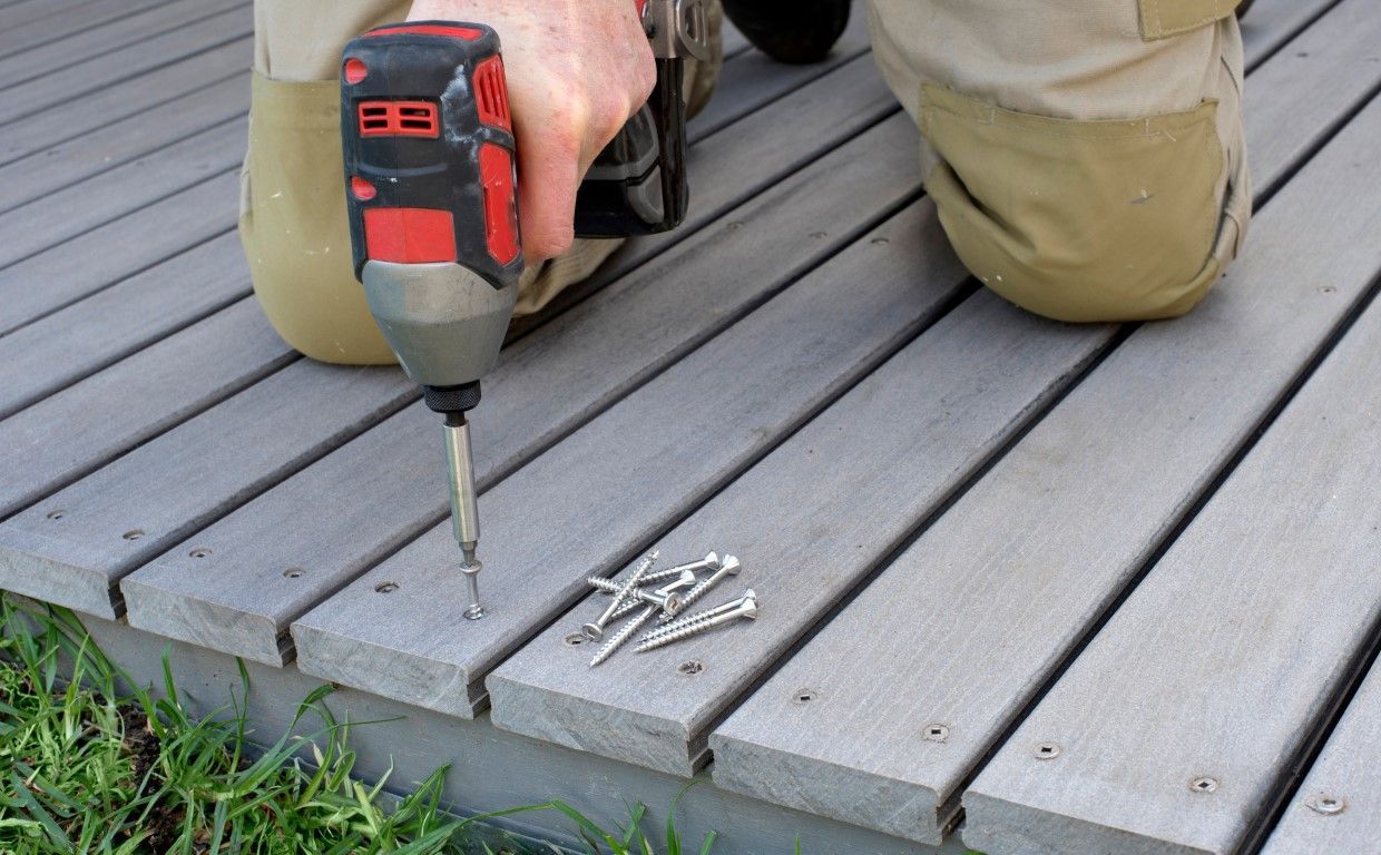 A person kneeling on a grey composite deck uses a cordless drill to secure boards, with a small pile of screws nearby.