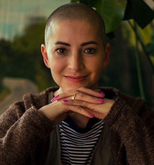 A woman with cancer and a shaved head is wearing a striped shirt and a brown jacket, smiling aftera free consultation with NYC Acupuncturist Marc Bystock