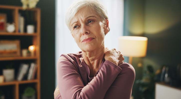 Elderly woman in a pink shirt clutches her shoulder, showing discomfort, in a well-lit living room.