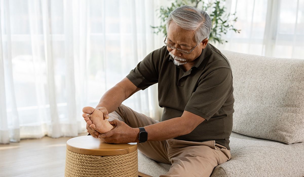 A man needing neuropathy treatments with acupuncture is seated on a sofa, massaging his foot with a small woven stool nearby.