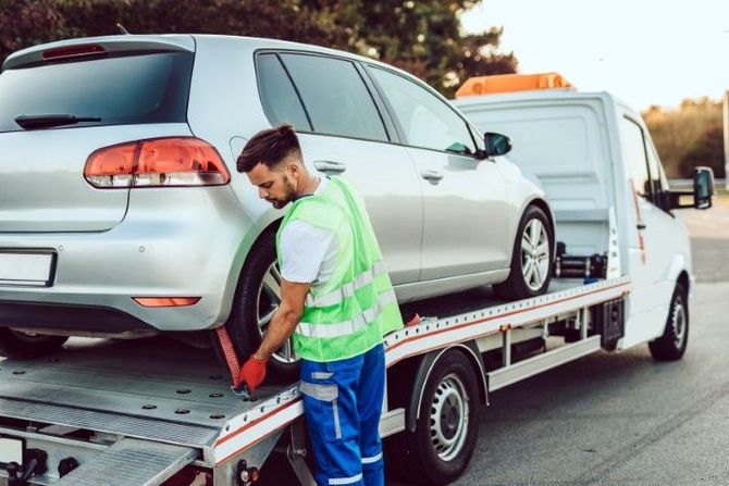 Tow truck worker loading a silver hatchback onto a flatbed on a roadside