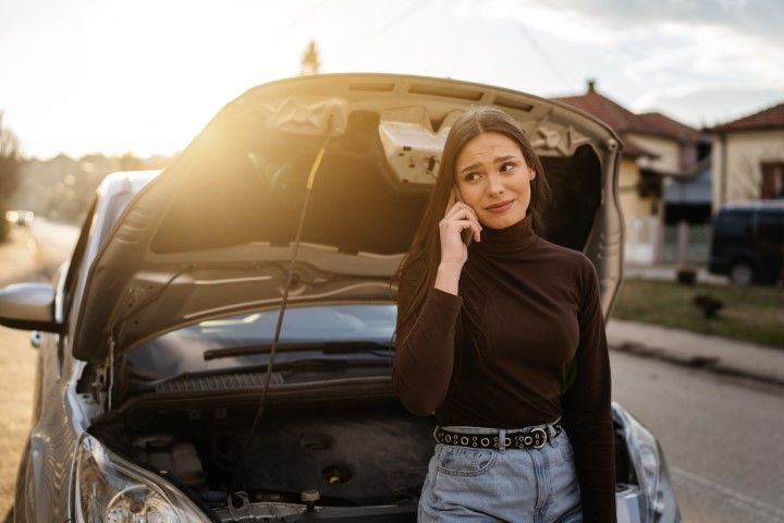 Woman standing by a black car with its hood open, talking on a phone at sunset.
