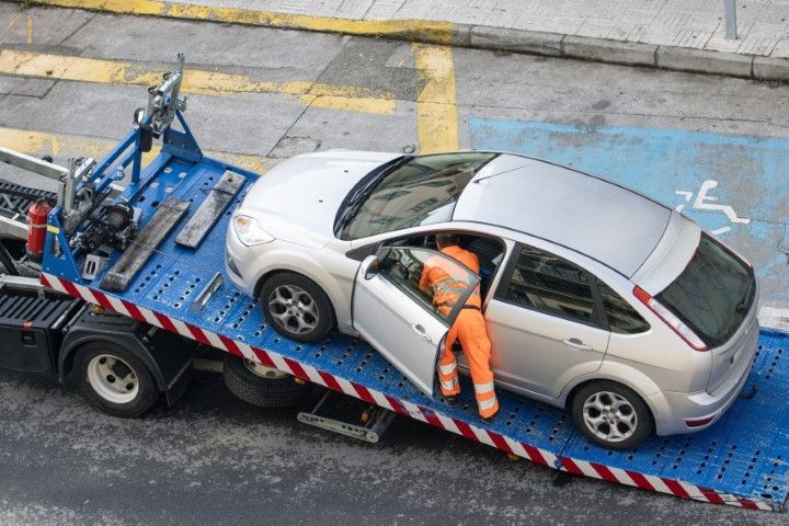 Tow truck operator in orange coveralls loads a silver car onto a flatbed truck