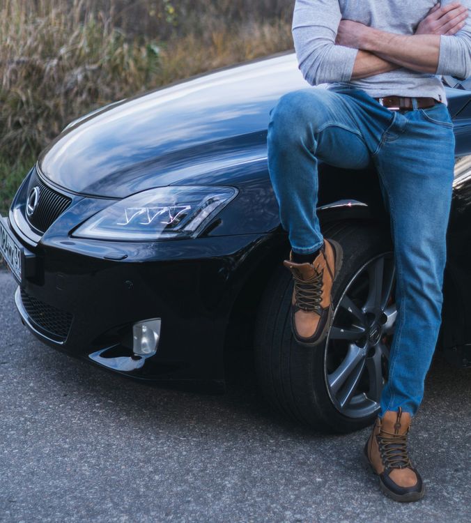Person leaning against a black sports car, wearing jeans and boots on a roadside.