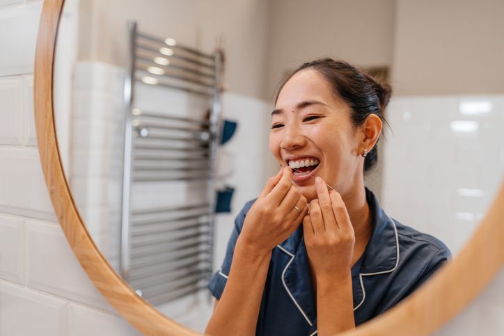 A person in pajamas smiles while flossing their teeth in front of a circular bathroom mirror.