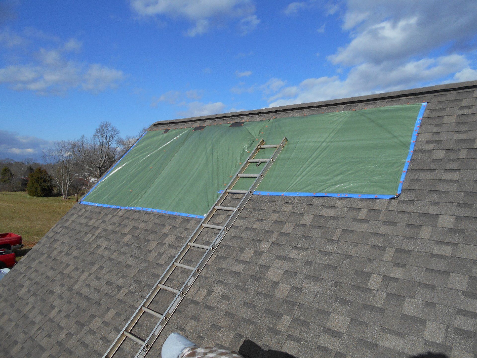 A tarp covers a portion of a roof. A ladder rests on the roof, and the sky is blue with clouds.